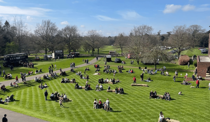A photo of a lush green lawn on a beautiful spring day. Families and crowds are dotted all over the grounds on blankets and having picnics.